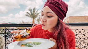 Frau mit rotem Kopftuch probiert ein süßes Gebäck auf Terrasse, mit Palme und Himmel im Hintergrund.