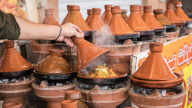 Mehrere Tajine-Töpfe auf Holzkohleöfen; eine Hand hebt den Deckel, während dampfendes Gemüsegericht sichtbar wird.