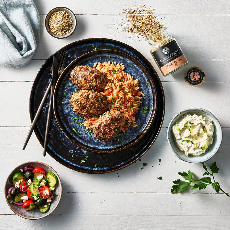Bifteki mit Reis auf Teller, dazu Dip, Salat und Gewürzglas, angerichtet auf hellem Tisch mit frischen Kräutern.