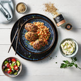 Bifteki mit Reis auf Teller, dazu Dip, Salat und Gewürzglas, angerichtet auf hellem Tisch mit frischen Kräutern.