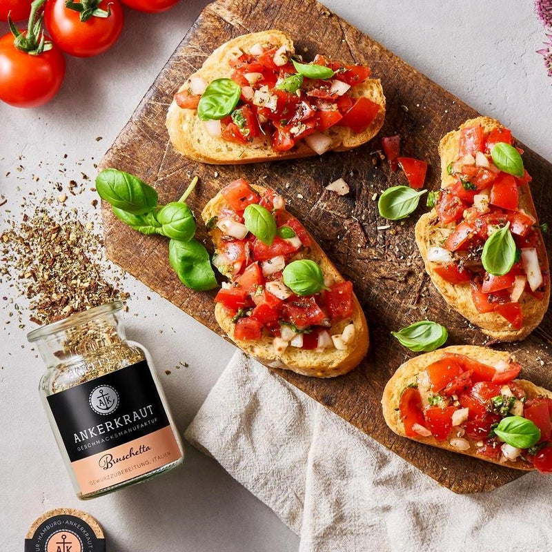 Bruschetta mit Tomaten und Basilikum auf Holzbrett, daneben Gewürzglas und frische Zutaten auf hellem Untergrund.