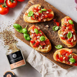 Bruschetta mit Tomaten und Basilikum auf Holzbrett, daneben Gewürzglas und frische Zutaten auf hellem Untergrund.