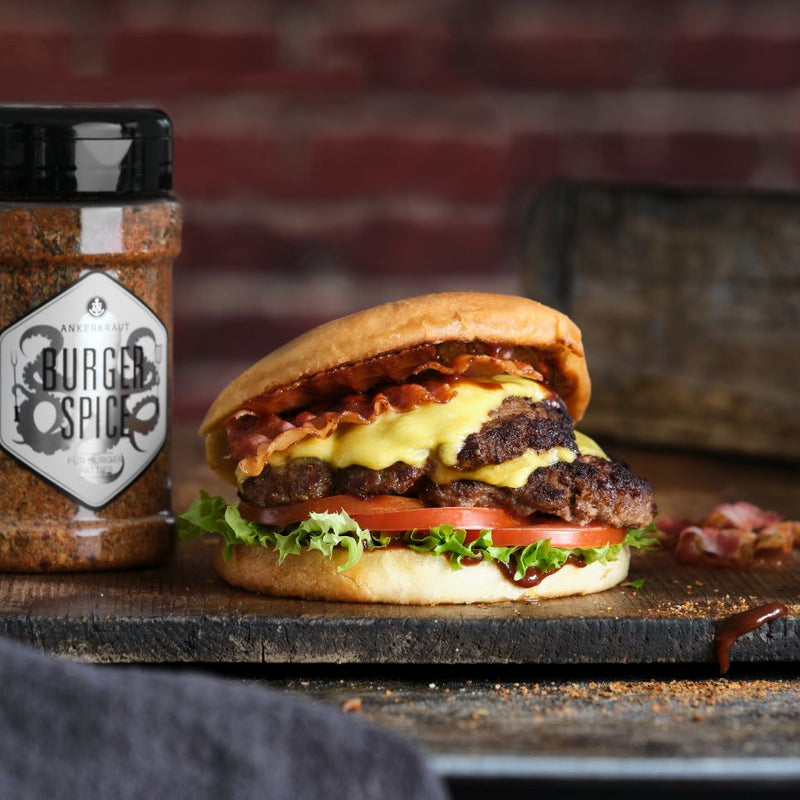 Saftiger Cheeseburger mit Bacon, Salat und Tomaten im Brötchen, daneben Burger-Gewürz auf rustikalem Holztisch.