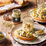 Eiersalat im Glas und auf Brot mit Schnittlauch, daneben Café de Paris Gewürzglas, Ei und Brot auf rustikalem Holztisch angerichtet.
