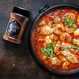 Cajun-Hähnchenpfanne mit Paprika in würziger Sauce, garniert mit Kräutern, daneben Gewürzglas auf dunklem Holztisch.