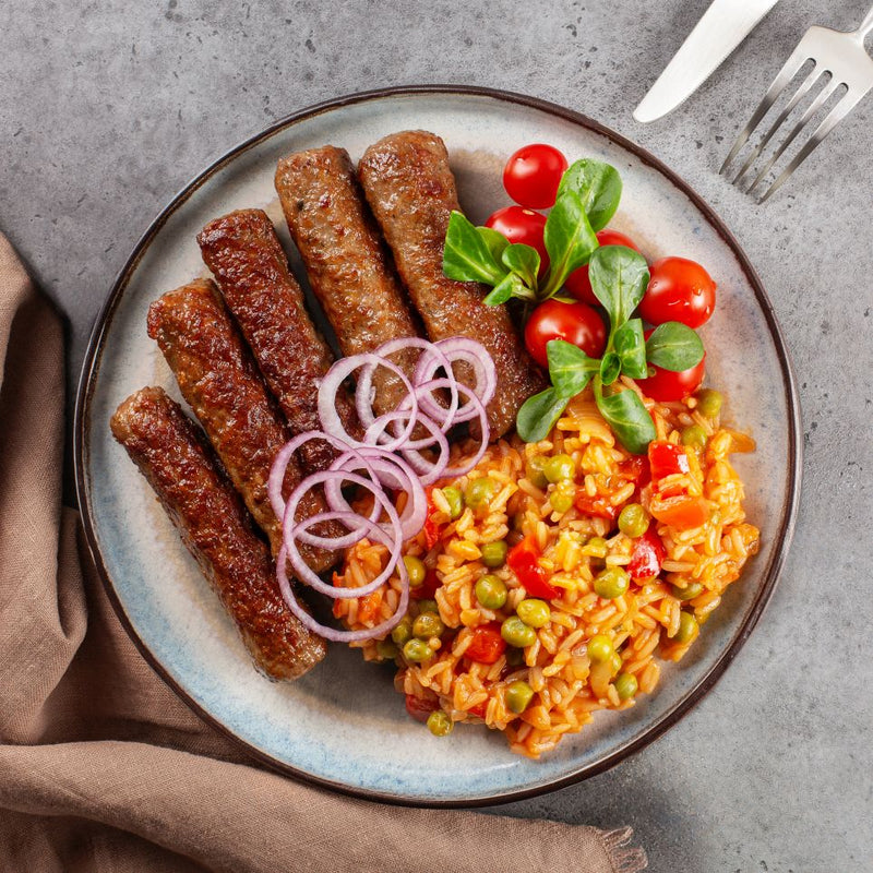 Cevapcici mit Djuvec-Reis, roten Zwiebeln und Tomaten, angerichtet auf Teller mit frischem Feldsalat.