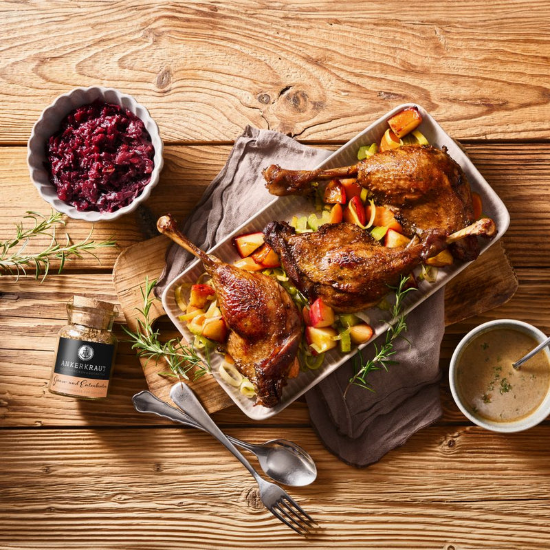Gebratene Entenschenkel auf Gemüse mit Apfelstücken, dazu Rotkohl, Sauce und Entengewürz auf rustikalem Holztisch.