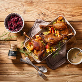 Gebratene Entenschenkel auf Gemüse mit Apfelstücken, dazu Rotkohl, Sauce und Entengewürz auf rustikalem Holztisch.