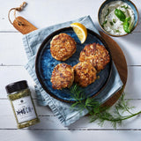 Fischfrikadellen mit Kräuterdip und Zitrone auf Teller, daneben Fischgewürz und frischer Dill auf hellem Tisch.
