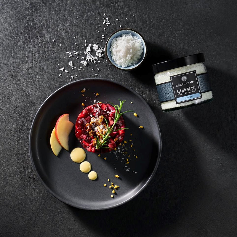 Rote Beete Tartar kunstvoll angerichtet mit Kräutern und Salz, daneben Schale mit Fleur de Sel und Gewürzglas auf dunklem Tisch.