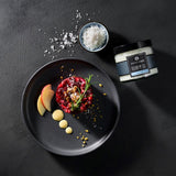 Rote Beete Tartar kunstvoll angerichtet mit Kräutern und Salz, daneben Schale mit Fleur de Sel und Gewürzglas auf dunklem Tisch.