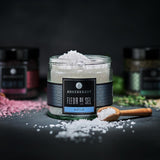 Glas mit Ankerkraut Fleur de Sel gefüllt, daneben Holzlöffel mit Salz, im Hintergrund unscharfe Gewürzgläser.