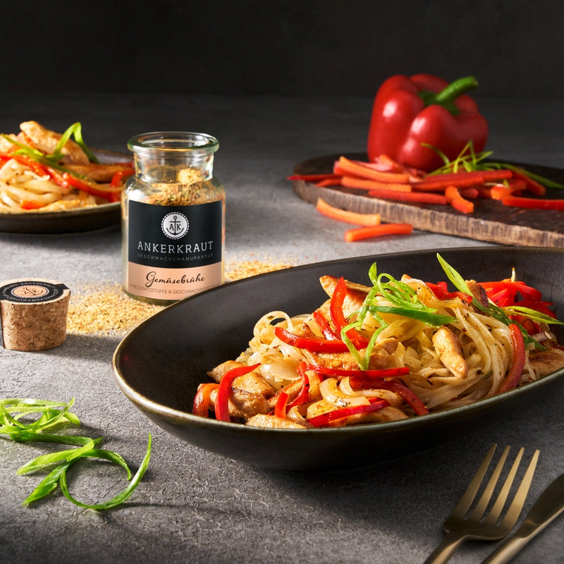 Nudelpfanne mit Hähnchenstreifen und Paprika in Schale, daneben Ankerkraut Gewürzglas und frisches Gemüse auf grauem Tisch.