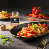 Nudelpfanne mit Hähnchenstreifen und Paprika in Schale, daneben Ankerkraut Gewürzglas und frisches Gemüse auf grauem Tisch.