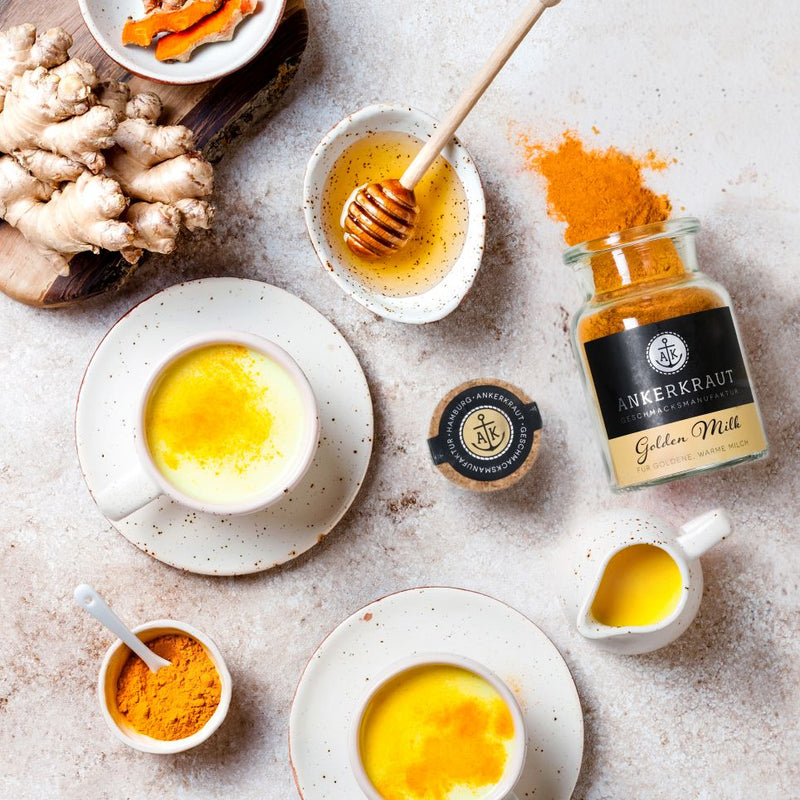Zwei Tassen Golden Milk mit Honig und Ingwer, daneben Gewürzglas, Kurkuma und frische Zutaten auf hellem Tisch.