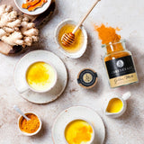 Zwei Tassen Golden Milk mit Honig und Ingwer, daneben Gewürzglas, Kurkuma und frische Zutaten auf hellem Tisch.