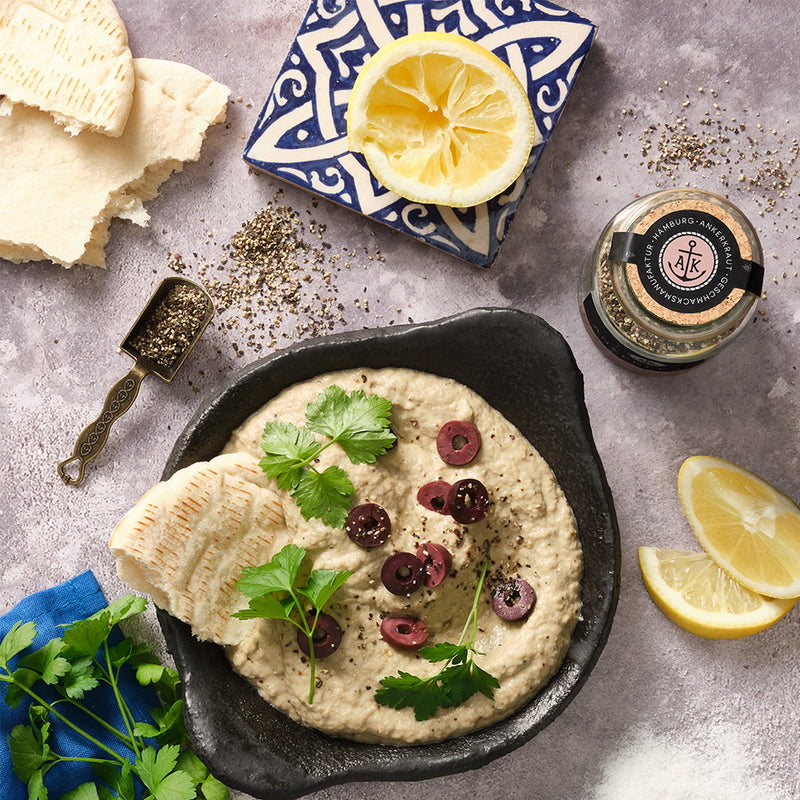 Cremiges Baba Ganoush mit Oliven und Petersilie, serviert mit Fladenbrot, Zitrone und Gewürzen auf hellem Untergrund.