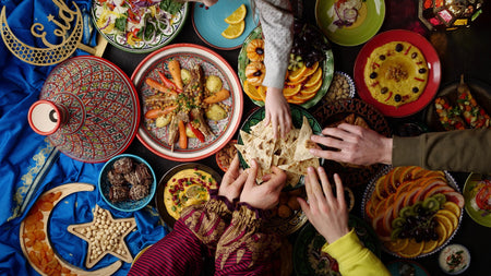 Bunt gedeckter Tisch mit Tajine, Fladenbrot, Obst und Mezze; mehrere Hände greifen gemeinsam nach Essen.