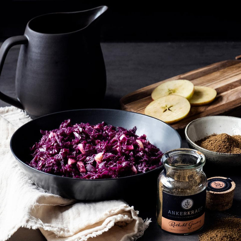 Rotkohl mit Apfelstücken in Schale, daneben Rotkohl Gewürz und frische Zutaten auf dunklem Tisch.