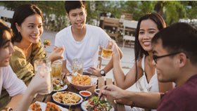 Gruppe junger Menschen sitzt draußen am Tisch, isst Snacks und stößt mit Getränken an.