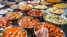 Große Auswahl an Street-Food-Gerichten in Metallschalen mit Saucen, Fleisch, Gemüse und Reis auf Marktstand.
