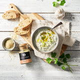 Cremiger Tzatziki-Dip mit Gurke und Olivenöl, dazu Brot, Knoblauch, Petersilie und Tzatziki-Gewürz auf hellem Tisch.