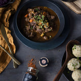 Wildragout in dunkler Sauce mit Gemüse auf tiefem Teller, dazu Kartoffelknödel und Wildgewürz auf dunklem Tisch.